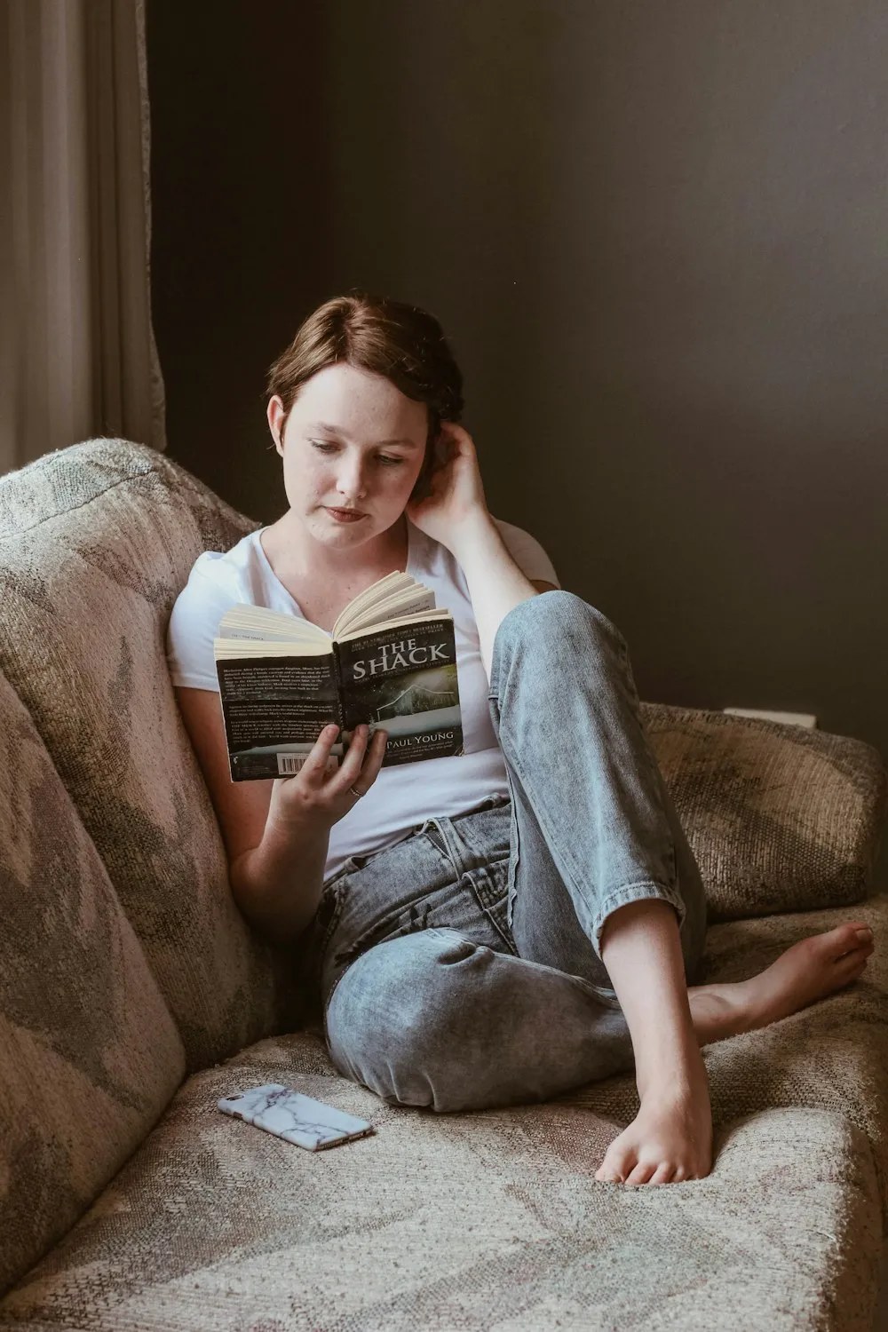 Woman reading a book on a sofa beside her phone